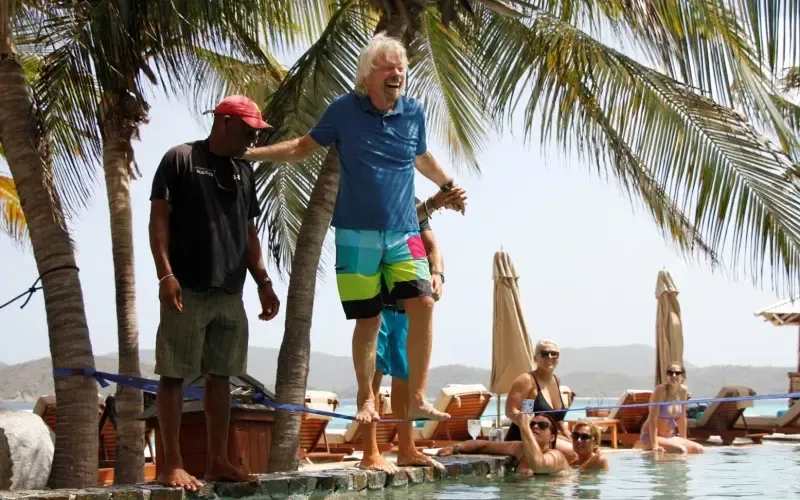 Richard Branson attempting to walk across a tightrope over a pool. Two people are helping him and others are watching on from the pool