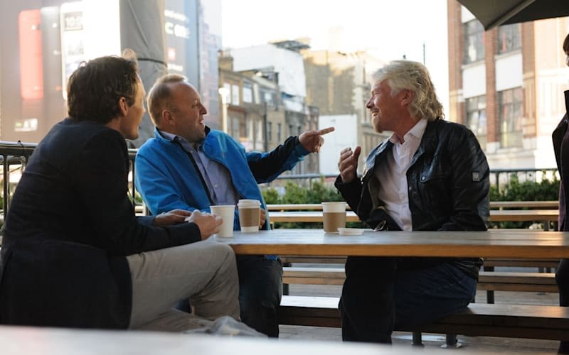 Richard Branson sitting at a picnic table talking and smiling with two men, one of whom is pointing at him