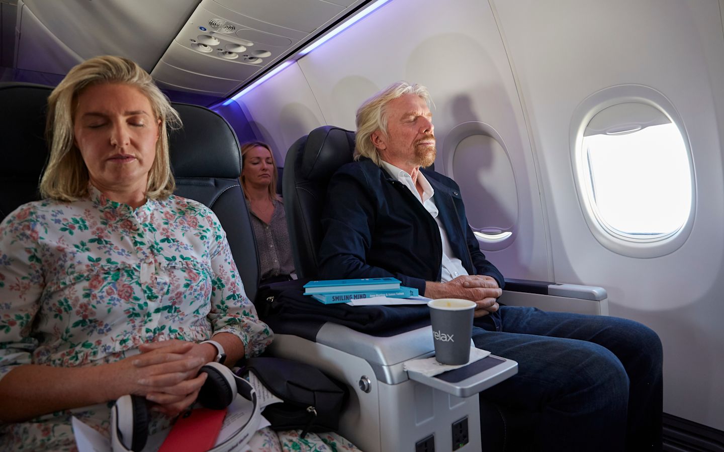 Richard Branson sitting on a plane, with his eyes shut, meditating. There is a women next to him and in the seat behind, also meditating.