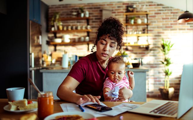 A mum tries to work at a kitchen table with a young child on her lap A mum tries to work at a kitchen table with a young child on her lap