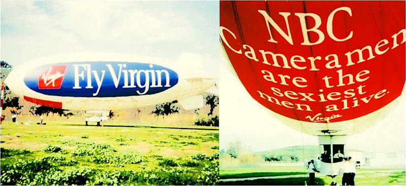 Virgin Blimp " Fly Virgin" in a field with the undercarriage of the blimp with "NBC Cameramen are the sexiest men alive" , the blimp has 4 persons surrounding it and 2 inside the gondola.