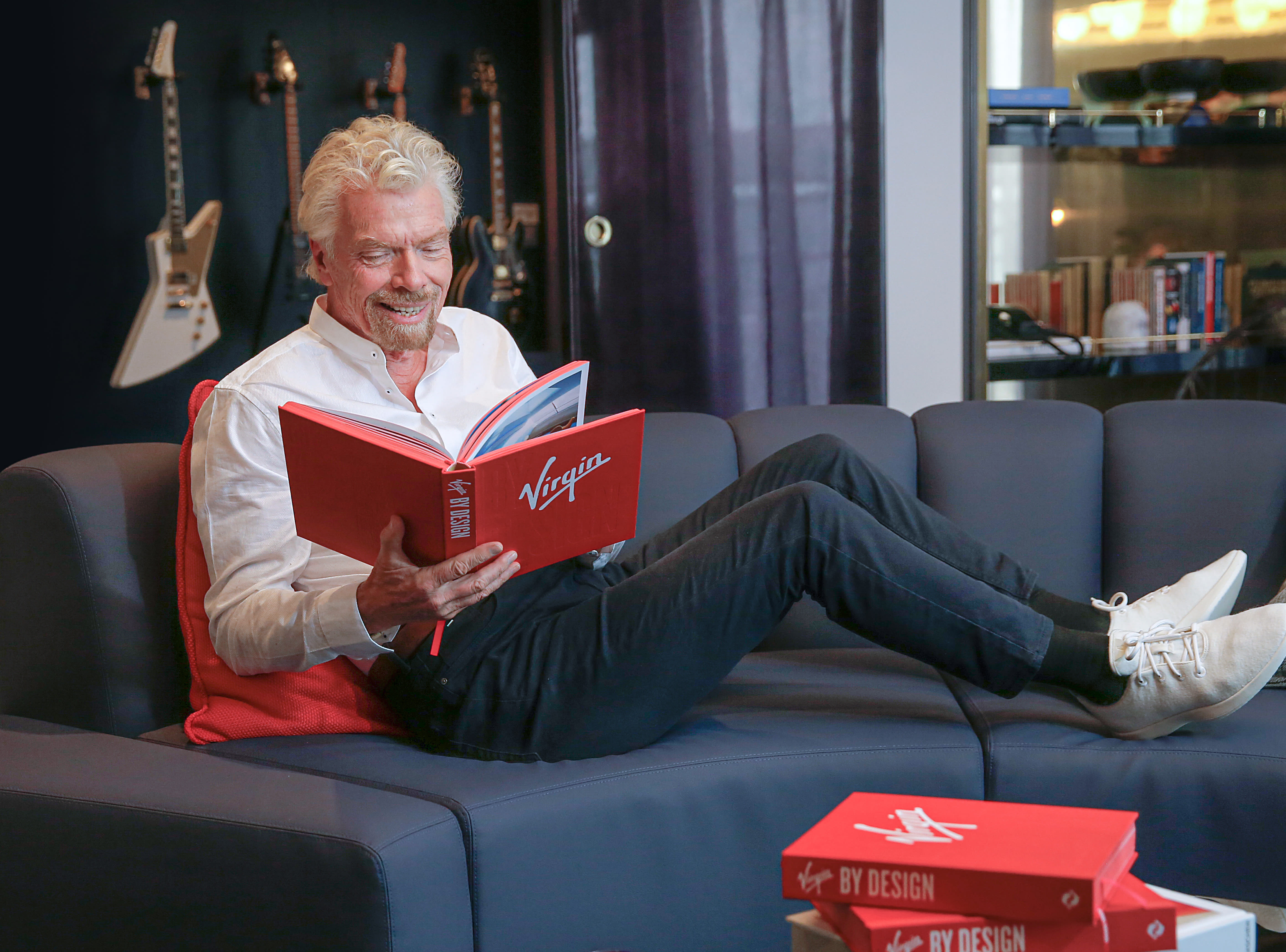Richard Branson relaxing on a sofa reading a copy of Virgin By Design