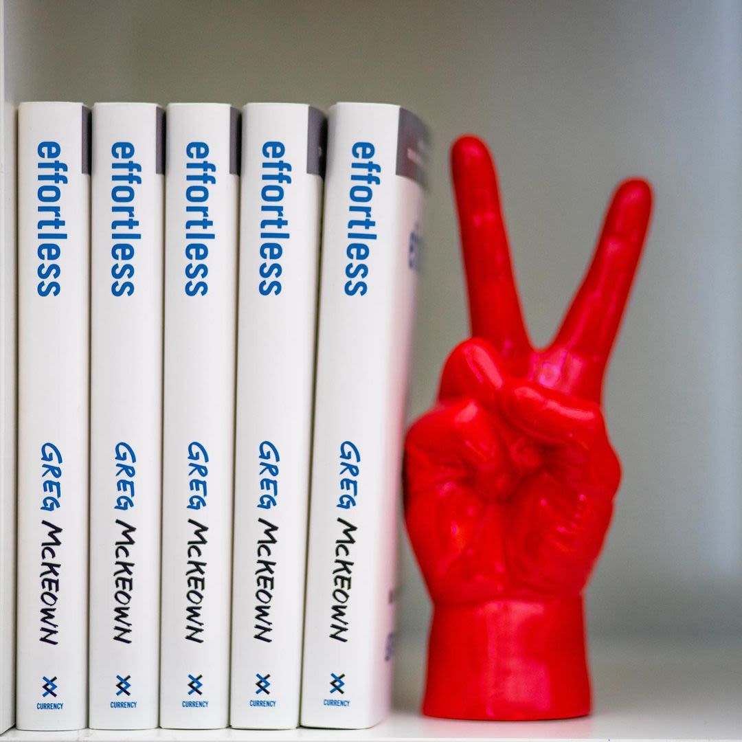 Five copies of Effortless by Greg McKeown on a book shelf next to a model of a hand showing a peace sign