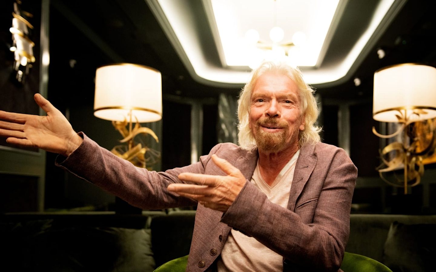 Richard Branson sitting inside a room on Virgin Voyages cruise ship smiling and gesticulating to the left