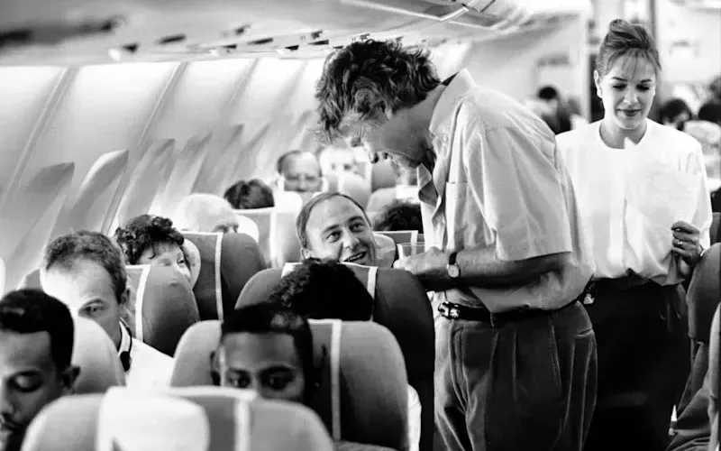 Black and white image of Richard Branson inside an aeroplane talking to a seated customer 