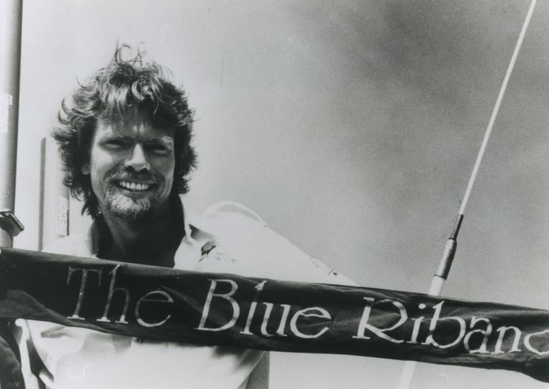 A black and white image of Richard Branson smiling behind a Blue Riband banner