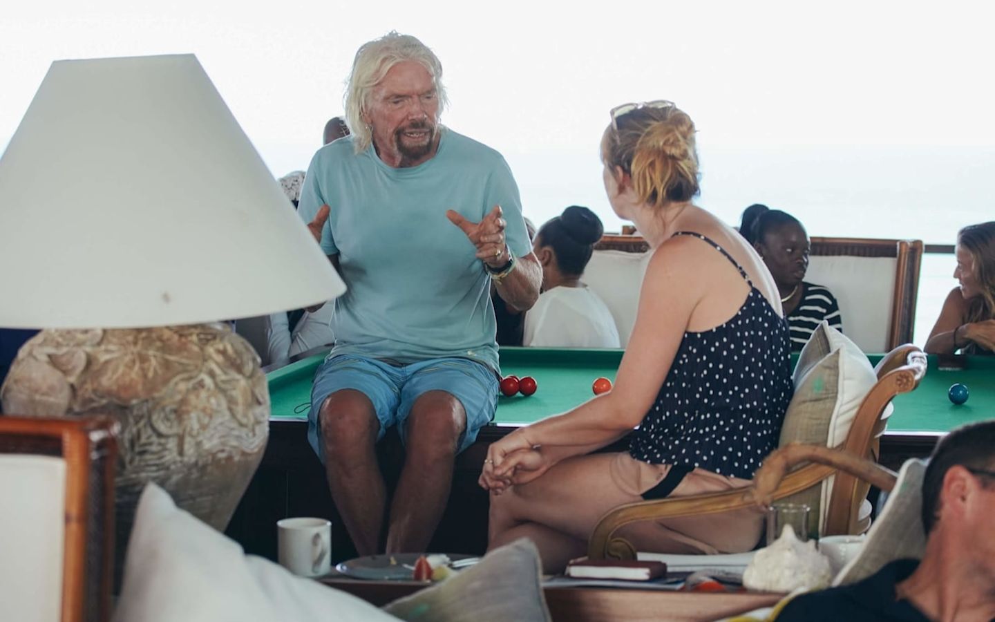 Richard Branson sitting on the edge of a pool table talking to a woman seated next to him