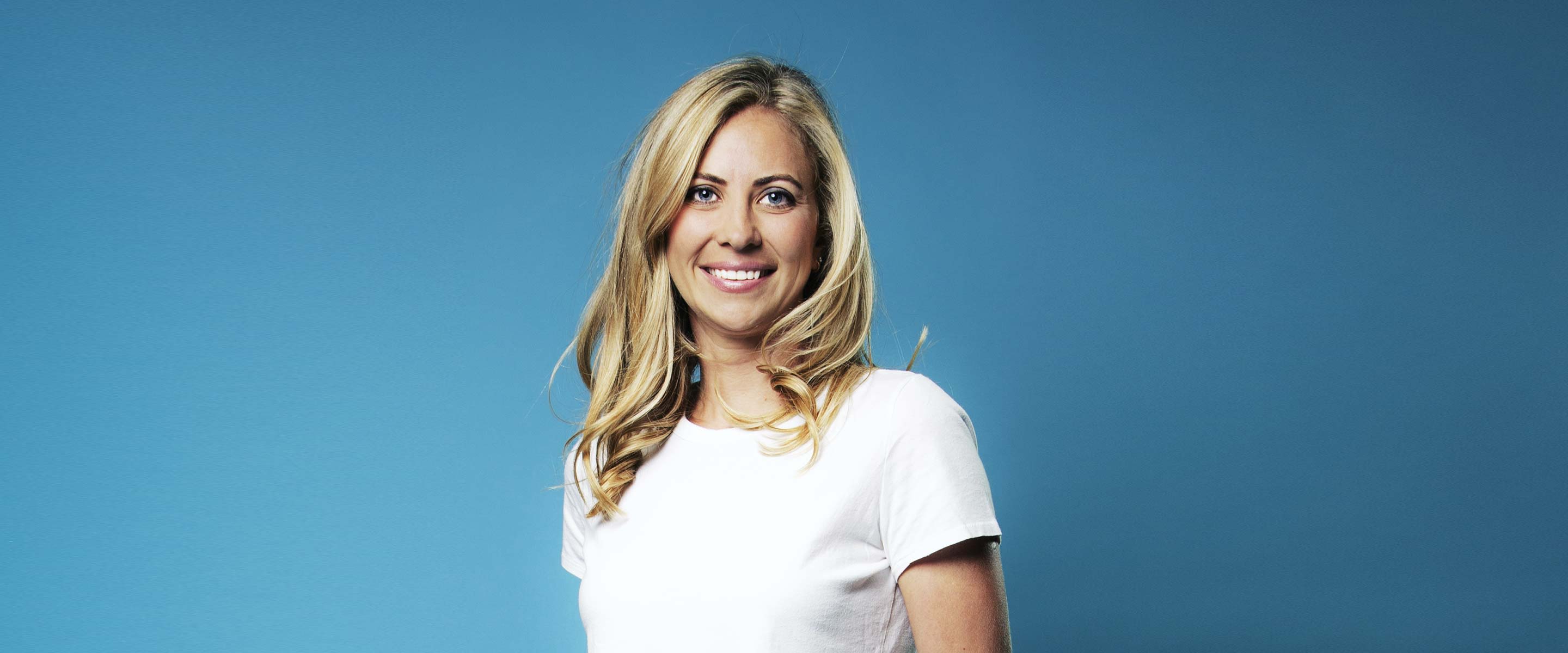 Holly Branson in a white t-shirt standing against a blue background and smiling into the camera