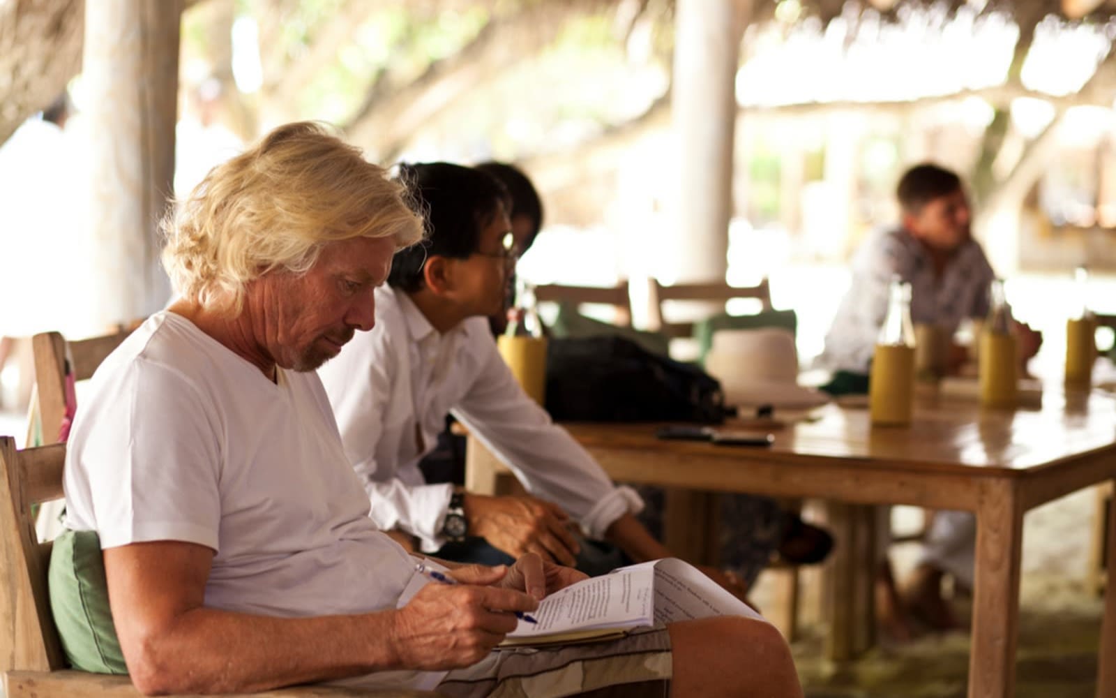 Richard Branson sitting in a chair writing, with people in the background