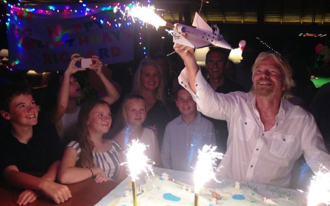 Richard with celebrating his birthday holding a sparkler up which is a model sized Virgin Galactic spaceship whilst he's surrounded by friends and family 