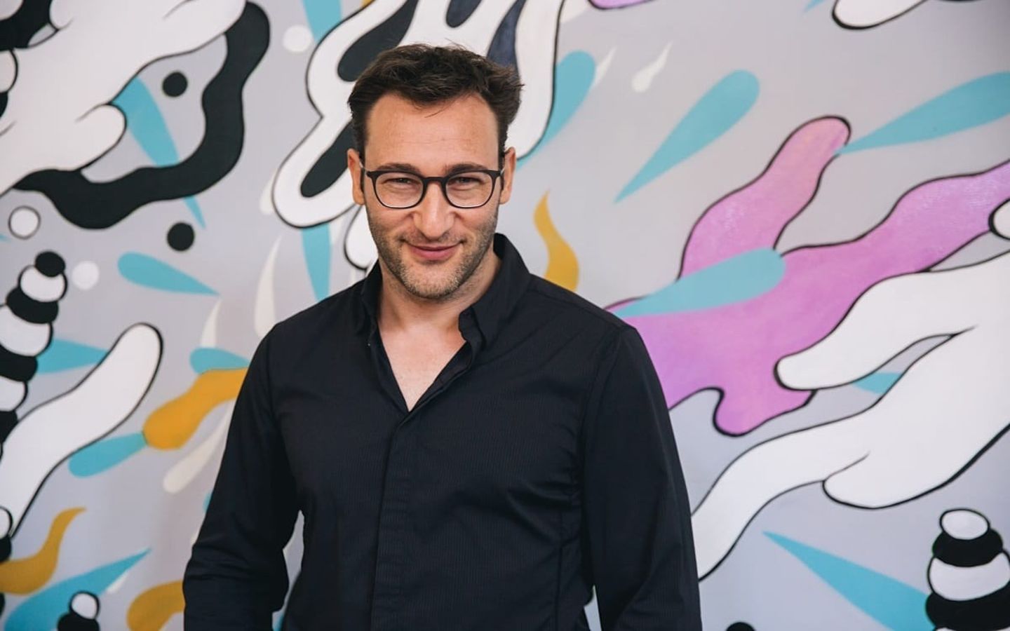 Simon Sinek. Wearing black, stands against a contemporary art background