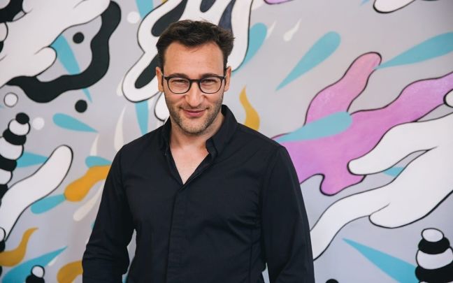 Simon Sinek.  Wearing black, stands against a contemporary art background