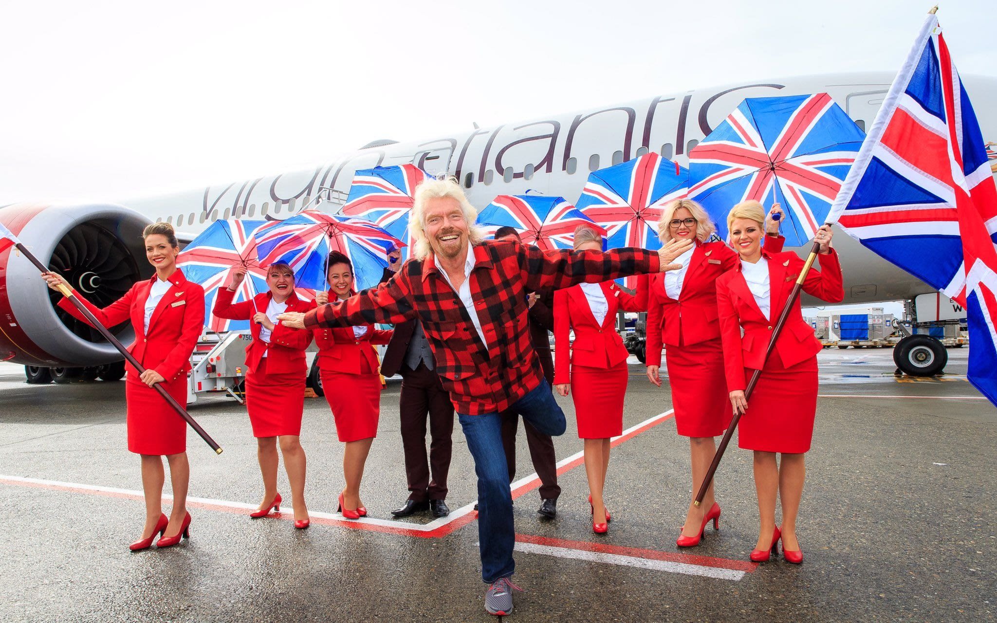 Richard Branson stands in front of Virgin Atlantic cabin crew holding Union Jack flags and umbrellas