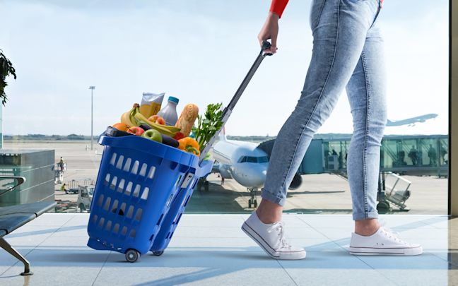 An image of a person wheeling a blue, filled Tesco shopping basket through an airport terminal. An image of a person wheeling a blue, filled Tesco shopping basket through an airport terminal.