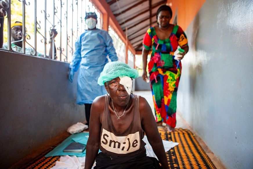 Nene sits up following trachoma surgery on the porch of her home while Forma,trachoma surgeon and his assistant Dionesa clean up from the surgery | Sightsavers/Alyssa Mariner