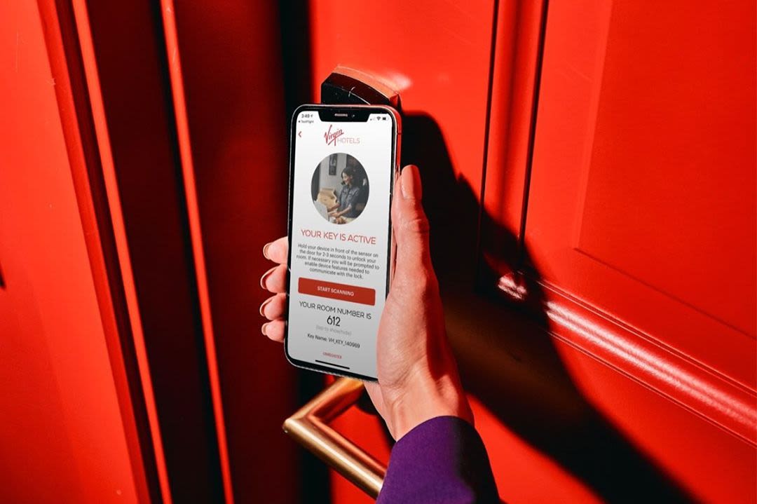 A hand holding an iPhone showing the Virgin Hotels app opening a red hotel room door
