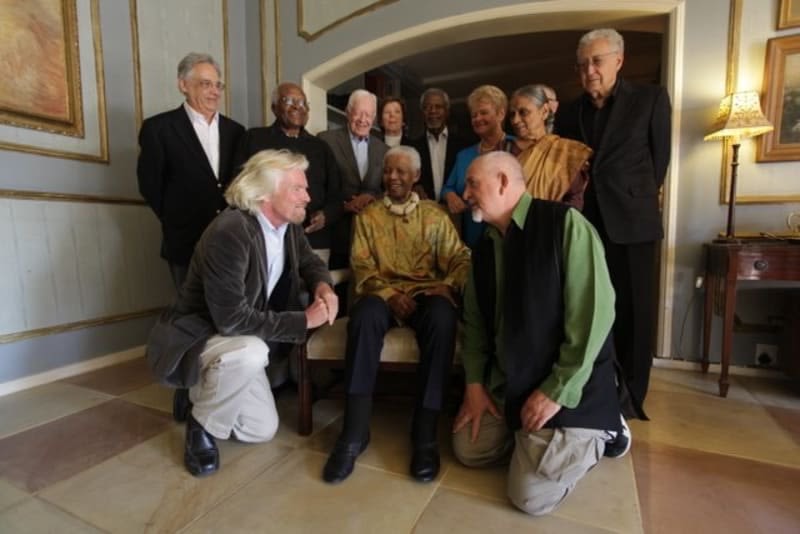 Richard Branson and Peter Gabriel kneel next to Nelson Mandela - surrounded by The Elders