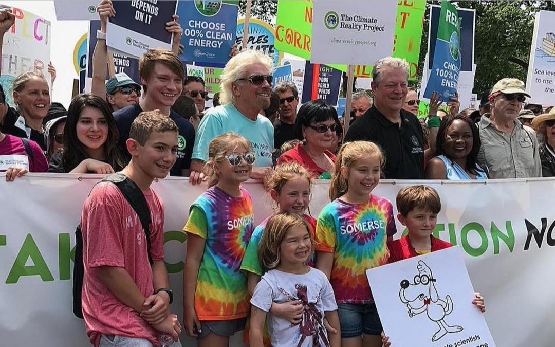 Richard Branson stands in solidarity at a Green Energy Protest.  He is smiling and surrounded by young, smiling protesters