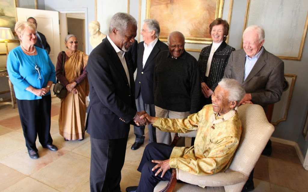 Nelson Mandela sat down on a chair shaking hands with Kofi Annan in front of a group of people.