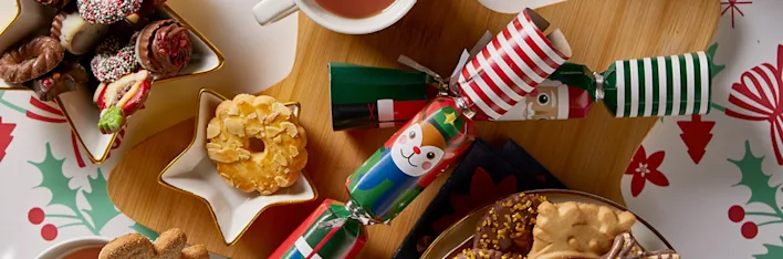 Feestelijke kerstsetting met thee, versierde koekjes in stervorm, kleurrijke crackers en een tafelkleed met hulstmotief.