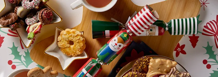 Feestelijke kerstsetting met thee, versierde koekjes in stervorm, kleurrijke crackers en een tafelkleed met hulstmotief.