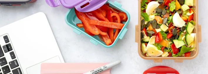 Werkplek met een laptop, een roze Sharpie-pen en twee lunchbakjes: één met gesneden rode paprika en één met een gemengde salade met onder andere gekookte eieren, avocado, pompoenpitten, rode paprika en bladgroenten.