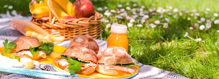 Een picknick met belegde broodjes, een klein flesje sinaasappelsap en een mand met bananen en een appel.