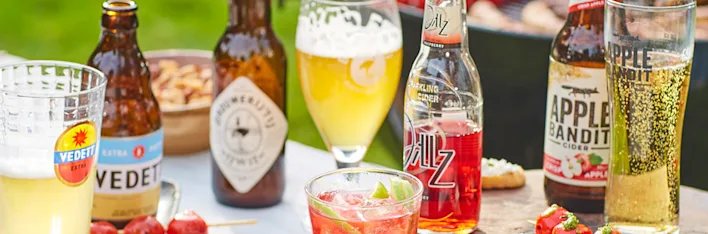 Diverse zomerse drankjes in flessen en glazen, waaronder bier, cider en een cocktail. Merken als Vedett, Apple Bandit en Jillz zijn zichtbaar.