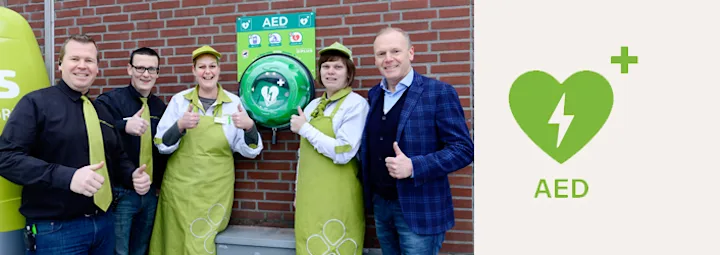 Groepsfoto met vijf PLUS-medewerkers voor een muur, bij een AED-apparaat.