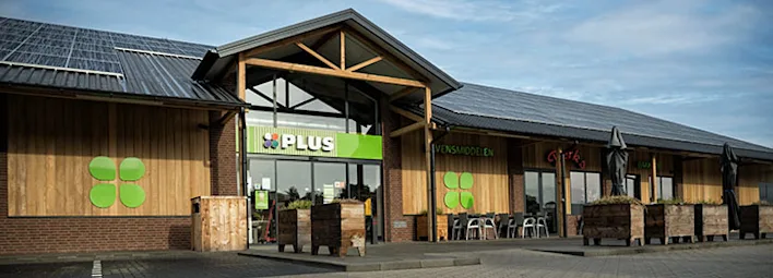 Vooraanzicht van een PLUS-supermarkt met een houten gevel en zonnepanelen op het dak.