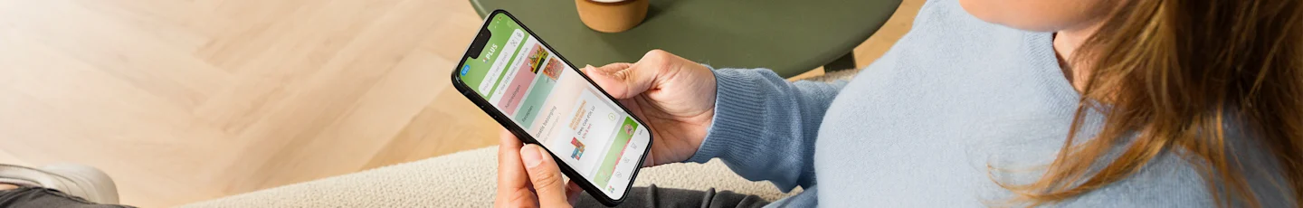 Een vrouw in een lichtblauwe trui zit op een bank en houdt een smartphone vast waarop een boodschappen-app geopend is, met aanbiedingen en producten zichtbaar op het scherm.