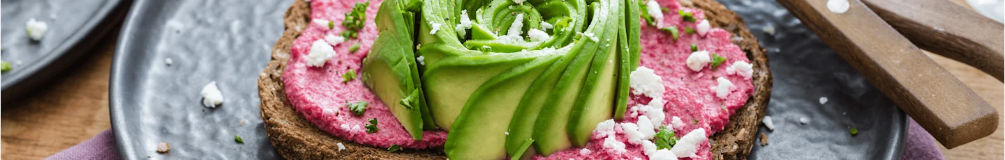 Een snee toast met bietenhummus, afgewerkt met een avocado-roos, witte kaas en groene kruiden.