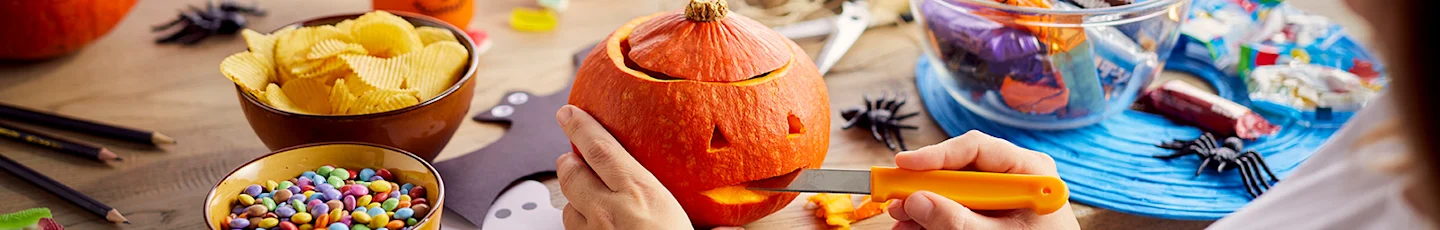 Persoon snijdt een pompoen met een oranje mes aan een tafel met Halloweenversiering, chips en snoep.