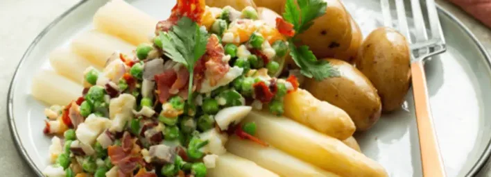 Wit bord met witte asperges, gegarneerd met een mix van erwtjes, spekblokjes en fijngesneden groenten en krieltjes.