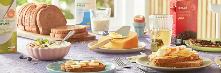 Ontbijttafel vol receptinspiratie, met onder andere een boterham met banaan, een boterham met gekookt ei, yoghurt met muesli en fruit, en diverse PLUS-producten zoals kaas, brood en chocoladehagelslag.