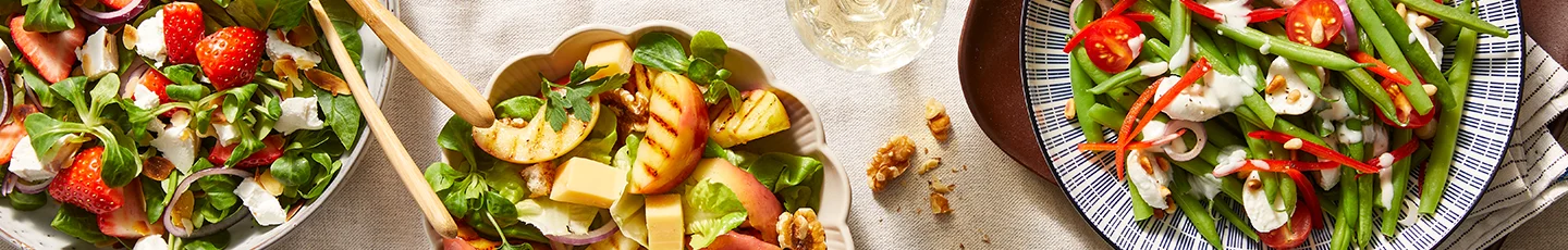 Zomerse saladerecepten van drie kleurrijke salades in kommen: één met aardbeien, feta en groene bladeren; een tweede met gegrilde perzik, walnoten en kaasblokjes; en een derde met sperziebonen, cherrytomaten en rode paprika, afgemaakt met romige dressing.