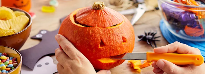 Persoon snijdt een pompoen met een oranje mes aan een tafel met Halloweenversiering, chips en snoep.