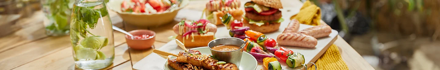 Zomerse gerechten, waaronder gegrilde spiesjes met vlees en groenten, sandwiches met diverse vullingen, een frisse salade in een kom en een sausdip in een schaal.