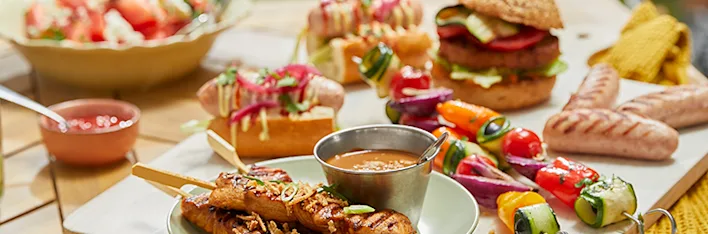 Zomerse gerechten, waaronder gegrilde spiesjes met vlees en groenten, sandwiches met diverse vullingen, een frisse salade in een kom en een sausdip in een schaal.