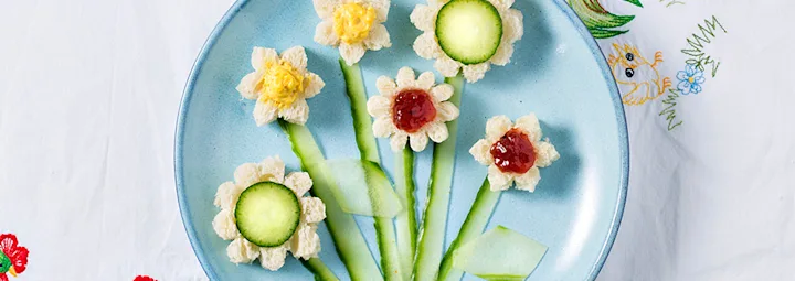 Ontbijt op een blauw bord: bloemen gemaakt van brood, komkommer en jam, met komkommerschijfjes als stelen en bladeren.
