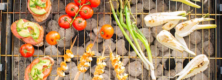 Gegrilde lekkernijen op een barbecue: links bruschetta met tomaat en avocado, in het midden spiesen met kip en kaas naast een tros cherrytomaten, rechts gegrilde bosui en venkel.