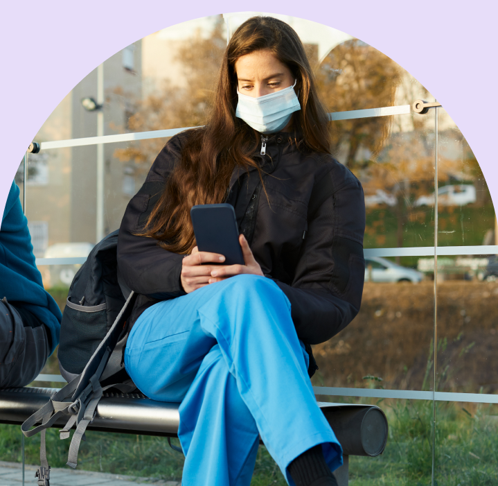 A girl wearing a mask sitting cross legged on a bench outside and using her phone