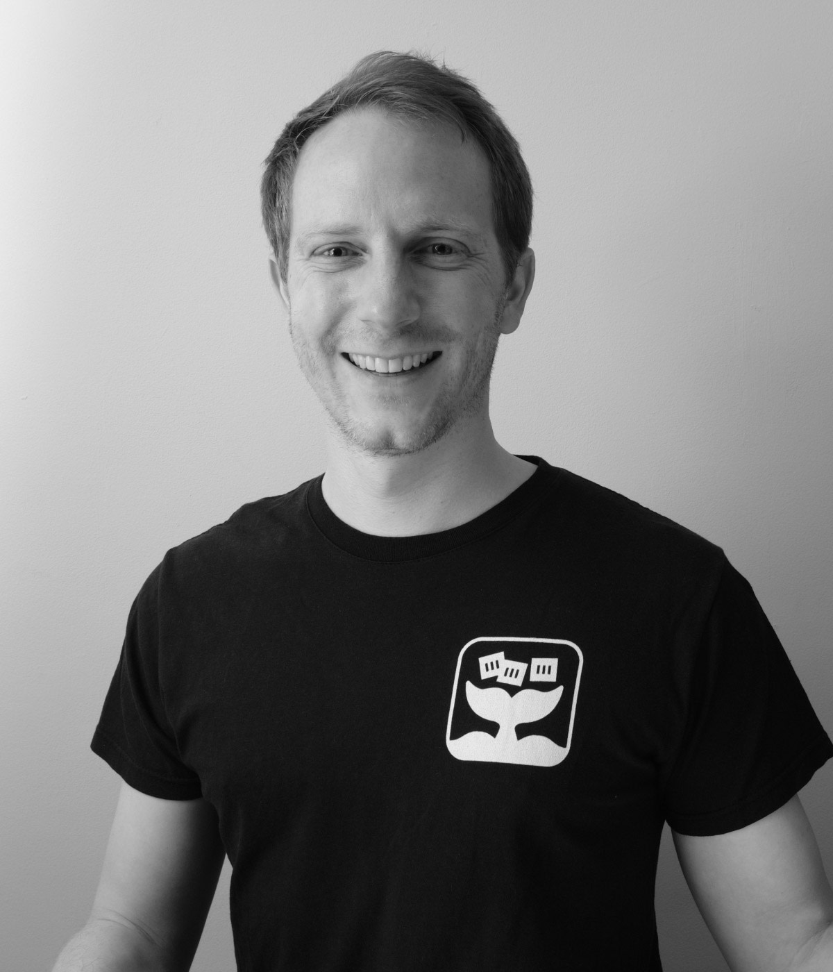 Black and white photo of Alex Ellis smiling, wearing an OpenFaaS branded t-shirt.