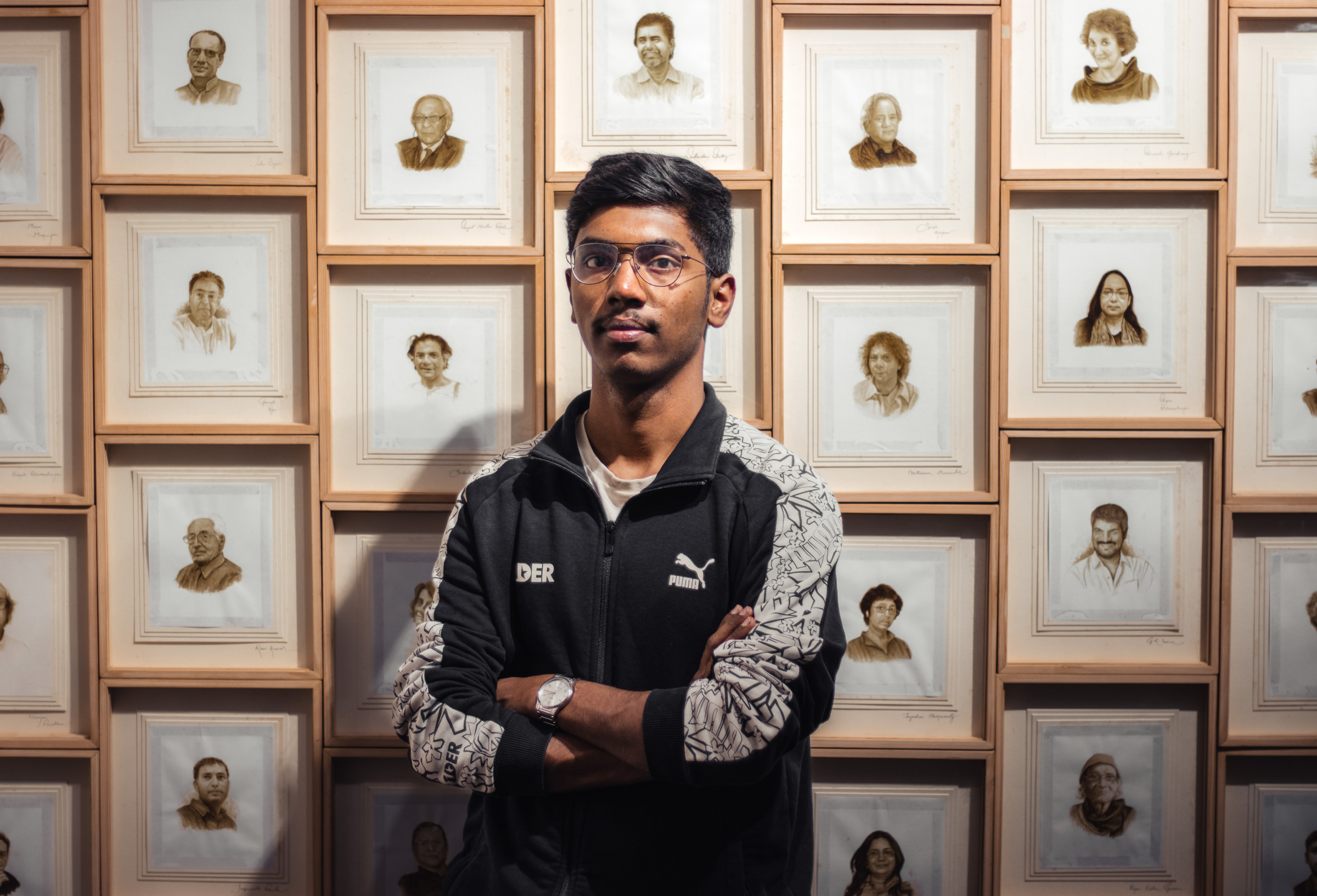 Author Rohan in a black-and-white Puma jacket with his arms crossed standing in front of a gallery wall of portraits