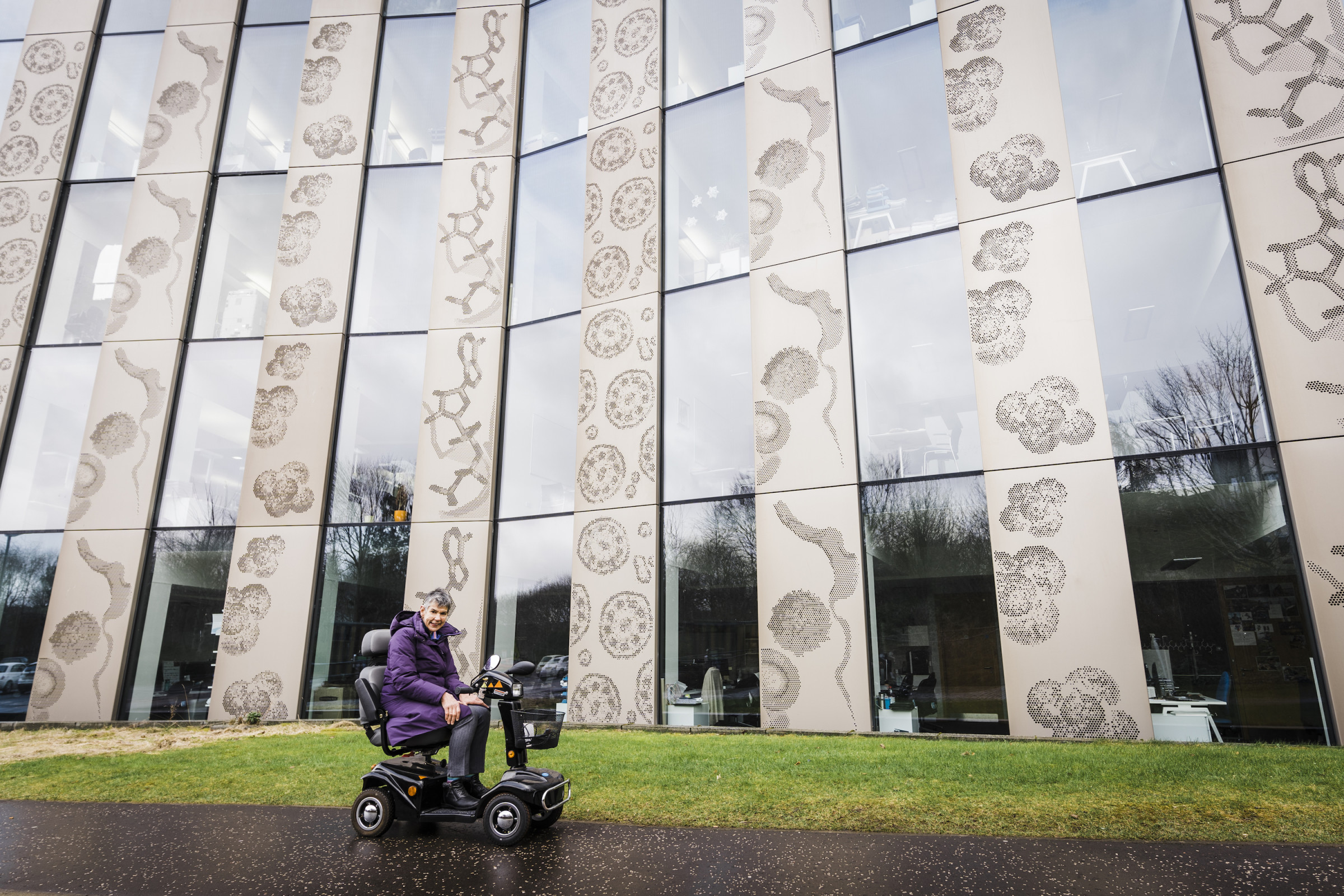 In a purple winter coat, Annalu sits in her motorized bicycle in front of a large building on the University of Dundee campus.