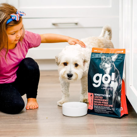Young girl petting dog beside bag of GO! SOLUTIONS SENSITIVITIES Limited Ingredient Grain-Free Salmon Recipe dry food