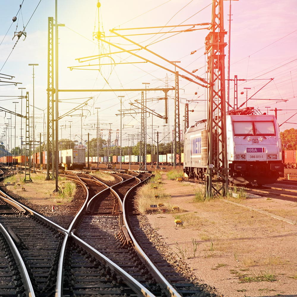 Das Foto zeigt Gleise samt Oberleitungen. Rechts steht ein Triebwagen. Im Hintergrund sind Waggons des Güterverkehrs zu sehen. 