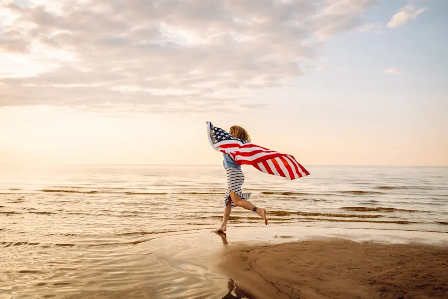 AFT 2025 Veterans Day Sale Hero. A joyful woman has fun walking along the beach with an American flag at sunset. A young woman is happy and enjoying nature with a US flag. Freedom and enjoyment concept.