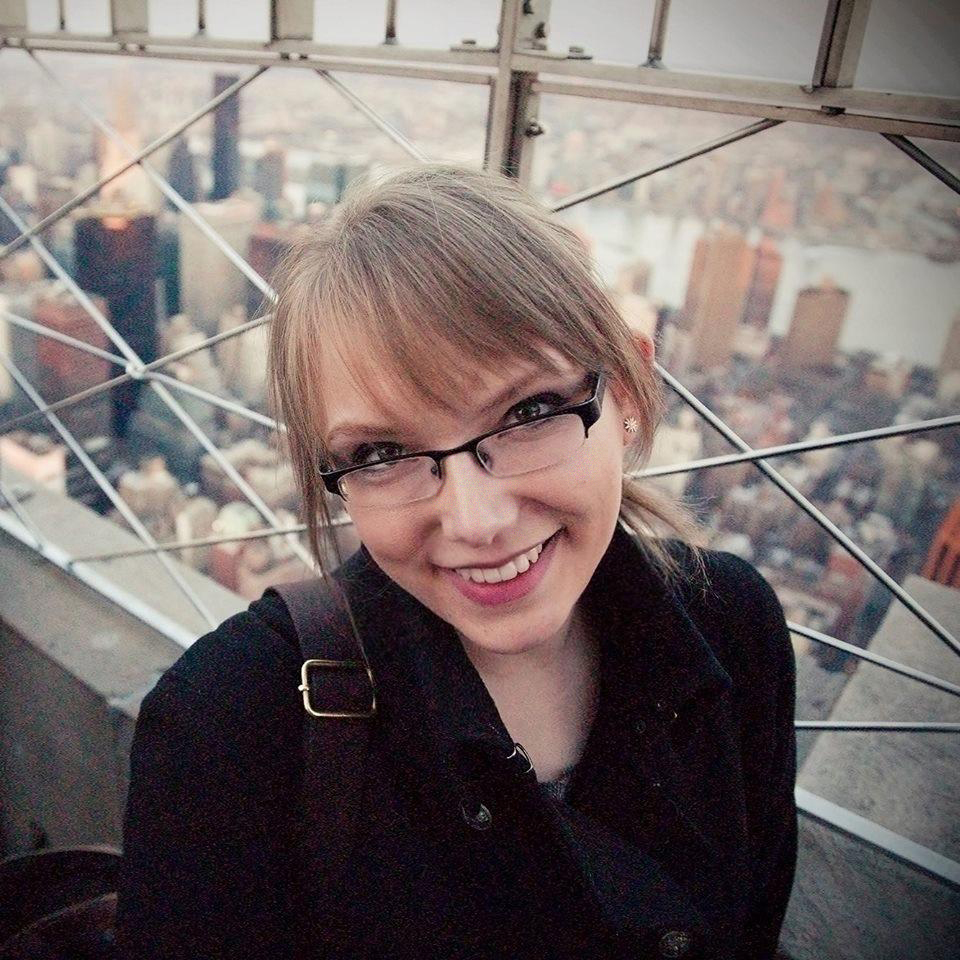 A white woman with blond hair and glasses standing and looking up with a cityscape in the background.}}