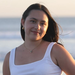 Headshot of a young woman with long dark hair. She wears a tank top with the ocean in the background.}}
